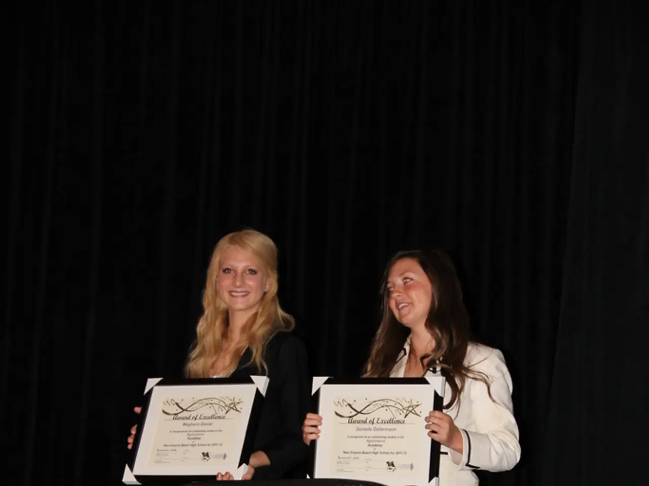 These two women are standing, smiling and holding certificates. In-front of them there is a table,...