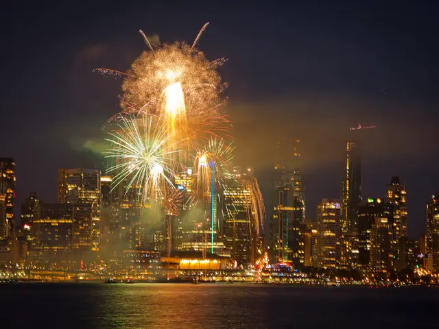 In the image there are buildings in the background with lights and there are crackers in the sky...