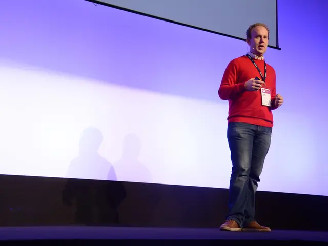 The man in the middle of the picture wearing the red T-shirt and ID card is standing. He is talking...