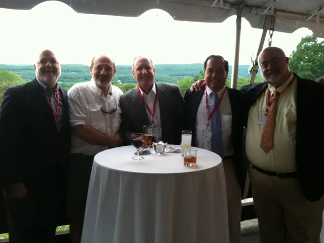 A group of five men are standing at a table with few glasses on it.