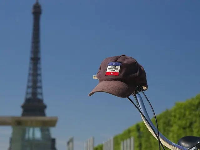 This image consists of a cap kept on the handle of a cycle. On the left, we can see a tower. On the...